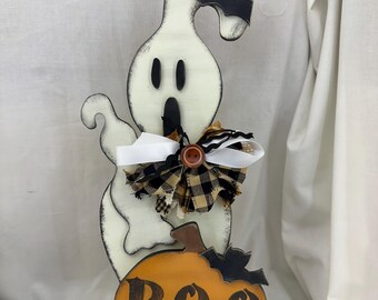 May include: A wooden Halloween decoration featuring a white ghost with black accents and a bat on top. The ghost holds an orange pumpkin with the word "BOO" in black. A decorative bow and button adorn the ghost's neck.