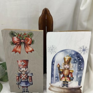 May include: Two wooden blocks with Christmas-themed illustrations. One features a nutcracker soldier and a red bow with holly. The other block shows a nutcracker inside a snow globe with snowflakes. The blocks are painted in neutral tones.