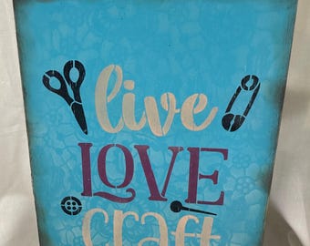 May include: A turquoise wooden sign with the words "live LOVE craft" in white and purple lettering. The sign features black scissors, a safety pin, a button, and a needle. The background has a floral pattern.