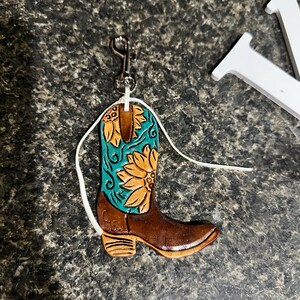 May include: A brown leather cowboy boot ornament with a turquoise shaft featuring a sunflower design. The boot has a silver-colored clip and a white string for hanging. The ornament is likely for a car or home decor.