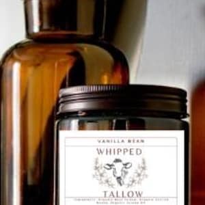 Op de afbeelding: Een glazen potkaars met een bruin deksel en een wit etiket. Het etiket zegt "Vanilla Bean Whipped" en heeft een koe-illustratie met het woord "Tallow" eronder. De kaars staat op een bruin oppervlak.