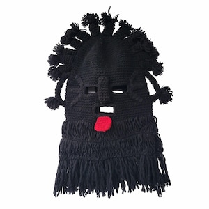 May include: Black knitted mask with a stylized face. The mask features a protruding nose, eye holes, and a red tongue. The top has multiple tufts, and the bottom has fringe.