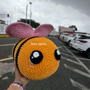 May include: A plush, round, yellow and black crocheted bee with large, expressive eyes and pink antennae. The bee has a smiling face and is held in a hand. The text "Brisa tejidos" is visible on the bee.