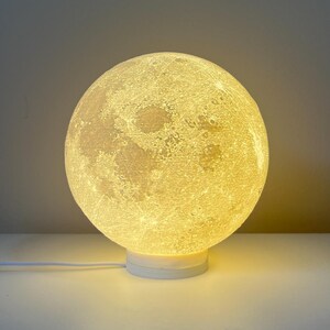 Pode incluir: Uma lâmpada branca em forma de lua com uma textura realista de superfície lunar. A lâmpada está acesa e emite um brilho amarelo quente. A lâmpada está sobre uma base branca e tem um cabo branco.