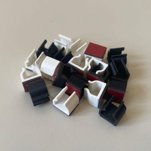 Pode incluir: Uma coleção de pequenos clipes de plástico em preto e branco, alguns com almofadas adesivas vermelhas. Os clipes têm um design em forma de U e são provavelmente usados para organizar ou fixar itens. A imagem mostra uma variedade de clipes.