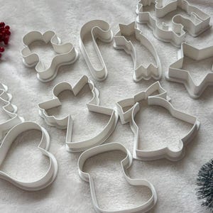 Può includere: Una collezione di formine per biscotti in plastica bianca a forma di elementi natalizi, tra cui bastoncino di zucchero, stella, campana, cuore e calzino. Le formine sono disposte su una superficie bianca.