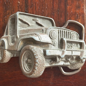 May include: A silver-colored metal belt buckle in the shape of a Jeep. The buckle depicts the front of the vehicle, including the grill, headlights, and tires. The buckle is set against a dark wood background.