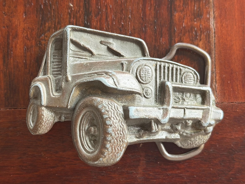 May include: A silver-colored metal belt buckle in the shape of a Jeep. The buckle depicts the front of the vehicle, including the grill, headlights, and tires. The buckle is set against a dark wood background.