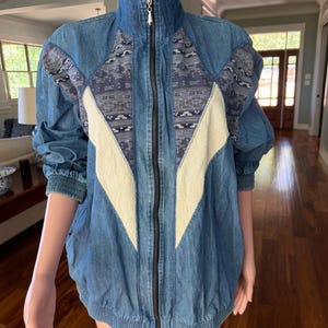 May include: A blue denim jacket featuring a high collar, a full-length front zipper, and elasticized cuffs and hem. The front panel displays contrasting cream-colored fabric inserts and dark blue patterned textile sections in a V-shaped design.