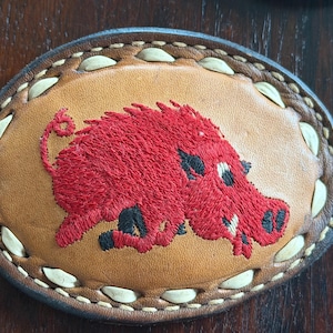 May include: Oval leather patch featuring a red embroidered Razorback hog. The hog has black accents and is centered on a tan background. The patch is bordered with a stitched design and a dark brown leather edge.