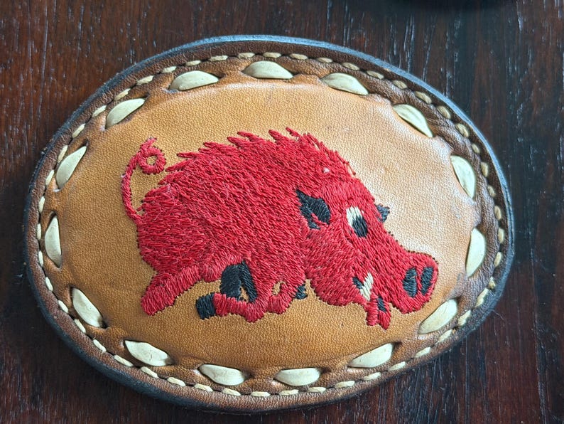 May include: Oval leather patch featuring a red embroidered Razorback hog. The hog has black accents and is centered on a tan background. The patch is bordered with a stitched design and a dark brown leather edge.