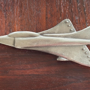May include: A silver-colored metal airplane figurine with a sleek design. The jet has a pointed nose, wings, and tail fins. The airplane is set against a dark brown wooden surface.