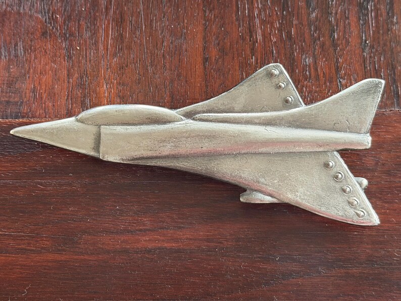 May include: A silver-colored metal airplane figurine with a sleek design. The jet has a pointed nose, wings, and tail fins. The airplane is set against a dark brown wooden surface.