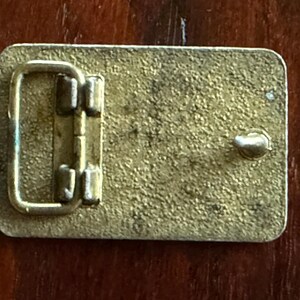 May include: Rectangular brass belt buckle with a textured surface. The buckle features a hinged bar and a single post for securing the belt. The metal has a worn, vintage appearance.