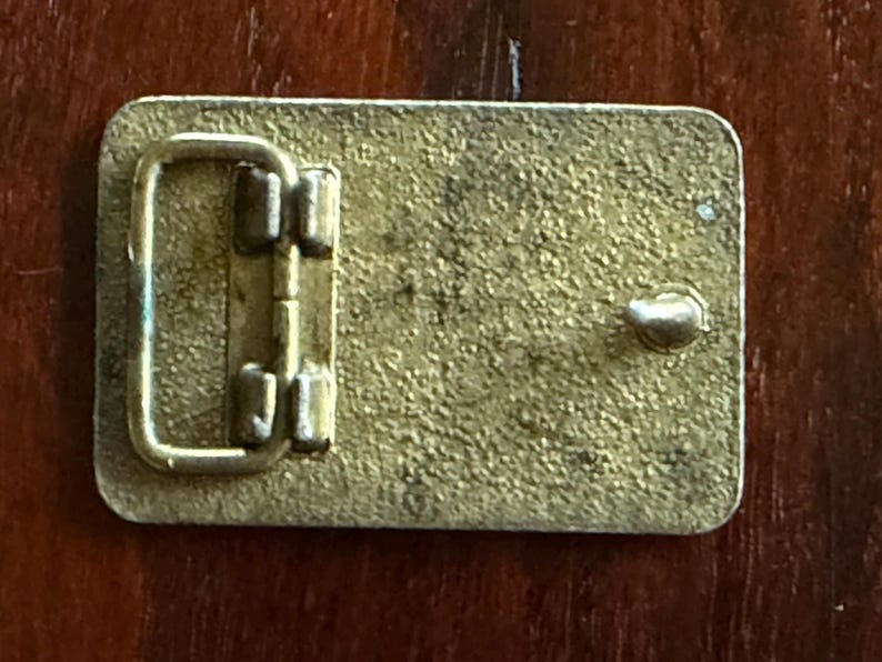 May include: Rectangular brass belt buckle with a textured surface. The buckle features a hinged bar and a single post for securing the belt. The metal has a worn, vintage appearance.