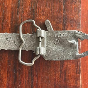 May include: A silver-colored metal belt buckle in the shape of a chainsaw. The buckle has a textured surface and the text "MBCL" is visible. The buckle is lying on a wooden surface.