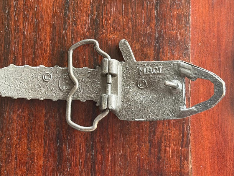 May include: A silver-colored metal belt buckle in the shape of a chainsaw. The buckle has a textured surface and the text "MBCL" is visible. The buckle is lying on a wooden surface.