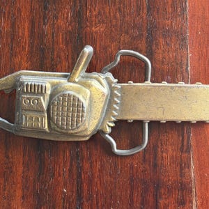 May include: A vintage-style belt buckle in the shape of a chainsaw. The buckle is made of a gold-toned metal with intricate details, including a handle, blade, and engine housing. The design is set against a dark wood grain background.