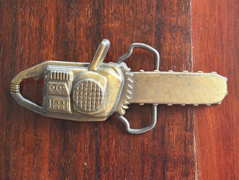 May include: A vintage-style belt buckle in the shape of a chainsaw. The buckle is made of a gold-toned metal with intricate details, including a handle, blade, and engine housing. The design is set against a dark wood grain background.
