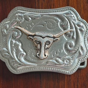 May include: Silver-toned metal belt buckle with a detailed floral and scroll design. The focal point is a gold-toned longhorn steer head. The buckle has a rectangular shape with a decorative edge, suitable for Western-themed accessories.