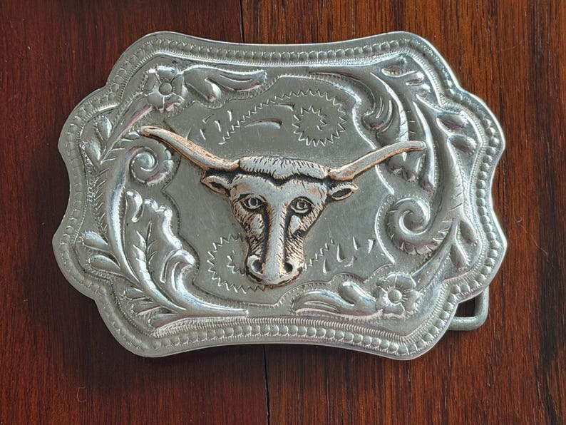 May include: Silver-toned metal belt buckle with a detailed floral and scroll design. The focal point is a gold-toned longhorn steer head. The buckle has a rectangular shape with a decorative edge, suitable for Western-themed accessories.
