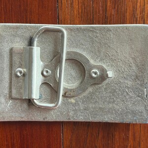 May include: A rectangular, silver-toned metal belt buckle with a loop and clasp mechanism. The buckle has a textured surface and is mounted on a wooden surface. The buckle's design is simple and functional, with visible screws securing the clasp.