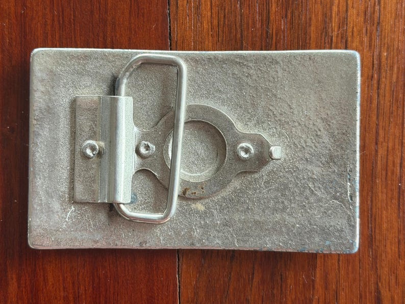 May include: A rectangular, silver-toned metal belt buckle with a loop and clasp mechanism. The buckle has a textured surface and is mounted on a wooden surface. The buckle's design is simple and functional, with visible screws securing the clasp.