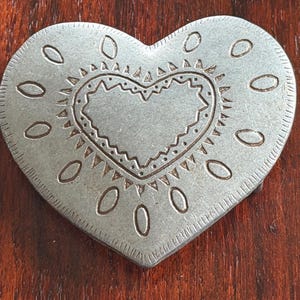 May include: A silver-colored heart-shaped belt buckle with an embossed design. The buckle features a heart within a heart, surrounded by oval shapes and a decorative border. The metal has a slightly aged appearance.