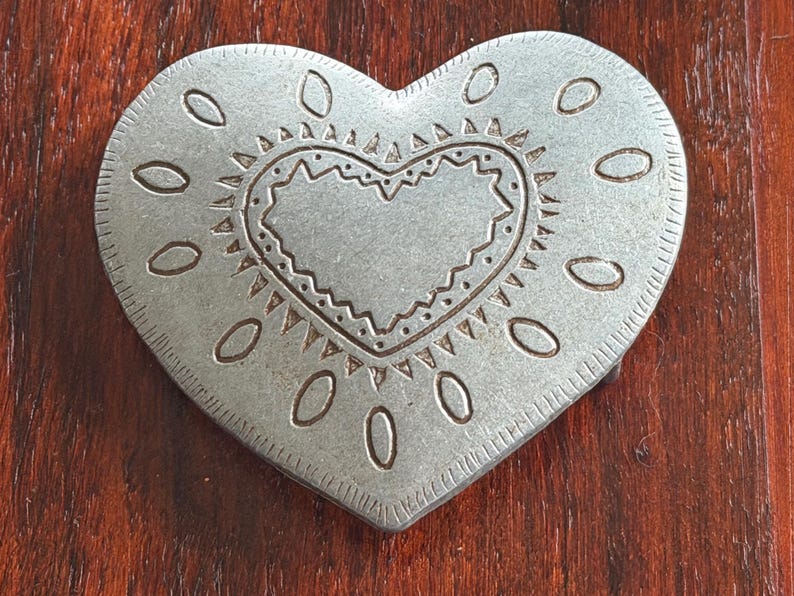 May include: A silver-colored heart-shaped belt buckle with an embossed design. The buckle features a heart within a heart, surrounded by oval shapes and a decorative border. The metal has a slightly aged appearance.