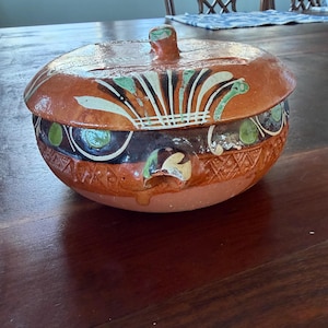 May include: A handcrafted, ceramic lidded bowl with a rustic, earthy aesthetic. The bowl features a warm, terracotta-colored base and lid, adorned with hand-painted designs in white, green, and black. The lid has a small handle. The bowl is likely for serving.