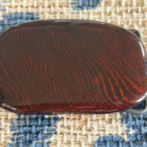 May include: A rectangular belt buckle with a glossy, dark reddish-brown surface. The buckle has a wood grain pattern and a silver-colored metal clasp.
