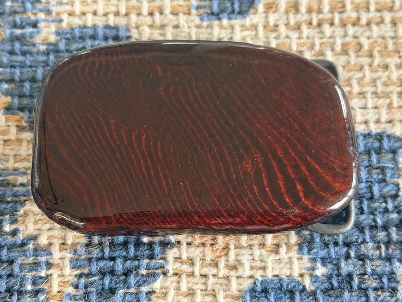 May include: A rectangular belt buckle with a glossy, dark reddish-brown surface. The buckle has a wood grain pattern and a silver-colored metal clasp.