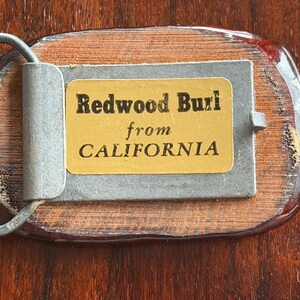 May include: A rectangular belt buckle with a wooden base and a metal clasp. The buckle features a label that reads "Redwood Burl from CALIFORNIA". The wood has a reddish-brown hue, and the metal components are silver.