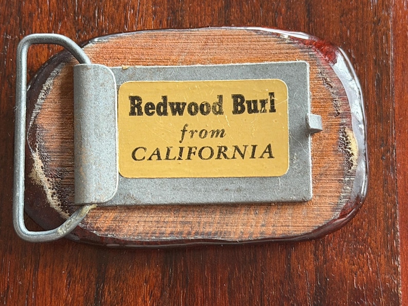 May include: A rectangular belt buckle with a wooden base and a metal clasp. The buckle features a label that reads "Redwood Burl from CALIFORNIA". The wood has a reddish-brown hue, and the metal components are silver.