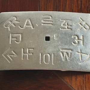 May include: A rectangular silver-colored metal belt buckle with engraved symbols and letters. The buckle has a slightly curved shape and a central square hole for attachment. The engraved designs include various symbols and the letters A, F, H, I, W, and Z.