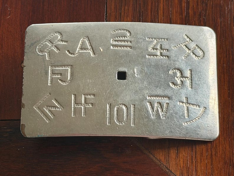 May include: A rectangular silver-colored metal belt buckle with engraved symbols and letters. The buckle has a slightly curved shape and a central square hole for attachment. The engraved designs include various symbols and the letters A, F, H, I, W, and Z.