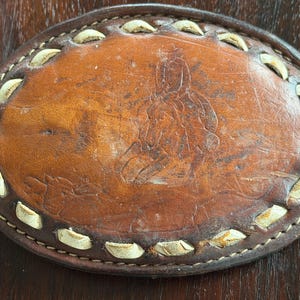 May include: Oval brown leather belt buckle with a stitched border and a decorative design. The buckle has a light brown border with white accents and a dark brown stitched edge. The center features an embossed image.