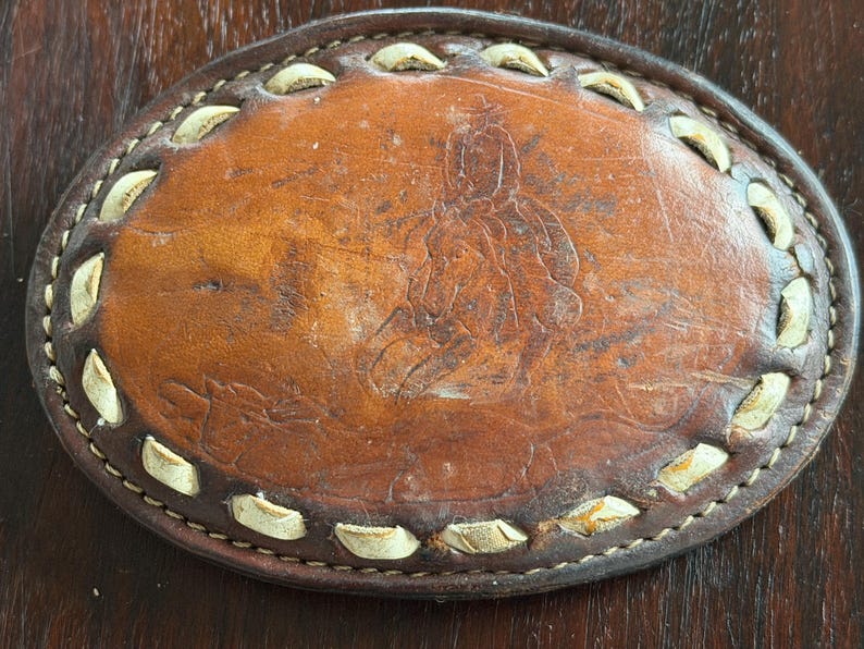 May include: Oval brown leather belt buckle with a stitched border and a decorative design. The buckle has a light brown border with white accents and a dark brown stitched edge. The center features an embossed image.