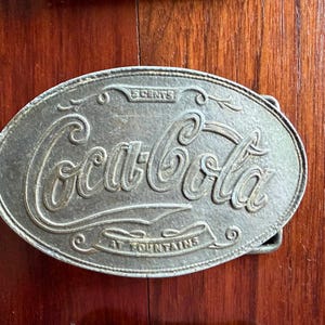 May include: Oval-shaped silver-colored belt buckle with the Coca-Cola logo in a cursive font. The buckle reads "5 CENTS" at the top and "AT FOUNTAINS" at the bottom. The buckle is made of metal and has a classic, vintage design.