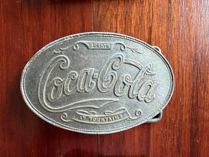 May include: Oval-shaped silver-colored belt buckle with the Coca-Cola logo in a cursive font. The buckle reads "5 CENTS" at the top and "AT FOUNTAINS" at the bottom. The buckle is made of metal and has a classic, vintage design.
