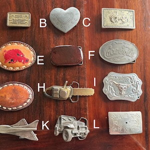 May include: A collection of belt buckles in various shapes and designs. Includes a rectangular Levi Strauss & Co. buckle, a heart-shaped buckle, and buckles with images of a rooster, a Coca-Cola logo, and a longhorn. Other buckles feature a chainsaw, a jet, and a jeep.