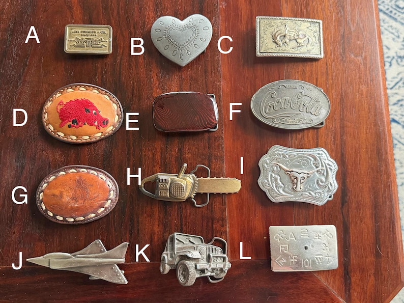 May include: A collection of belt buckles in various shapes and designs. Includes a rectangular Levi Strauss & Co. buckle, a heart-shaped buckle, and buckles with images of a rooster, a Coca-Cola logo, and a longhorn. Other buckles feature a chainsaw, a jet, and a jeep.