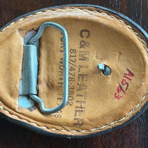 May include: Oval-shaped leather belt buckle with a metal clasp. The back of the buckle is tan leather with the text "C&M LEATHER" and "817/478-3802". The buckle has a stitched border and a red handwritten number "A1653".