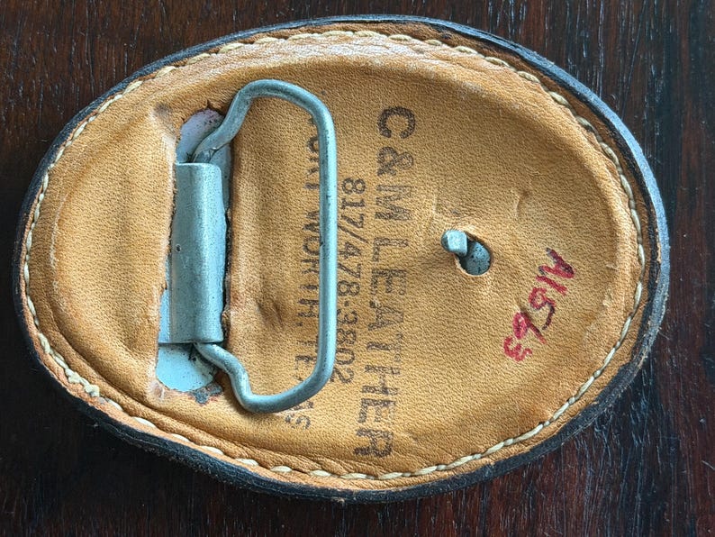 May include: Oval-shaped leather belt buckle with a metal clasp. The back of the buckle is tan leather with the text "C&M LEATHER" and "817/478-3802". The buckle has a stitched border and a red handwritten number "A1653".