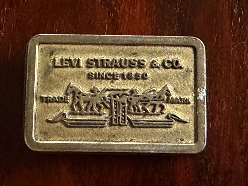 May include: A rectangular, gold-colored metal plaque with the words "LEVI STRAUSS & CO. SINCE 1850" and a graphic of two horses pulling a wagon. The words "TRADE" and "MARK" are also visible.