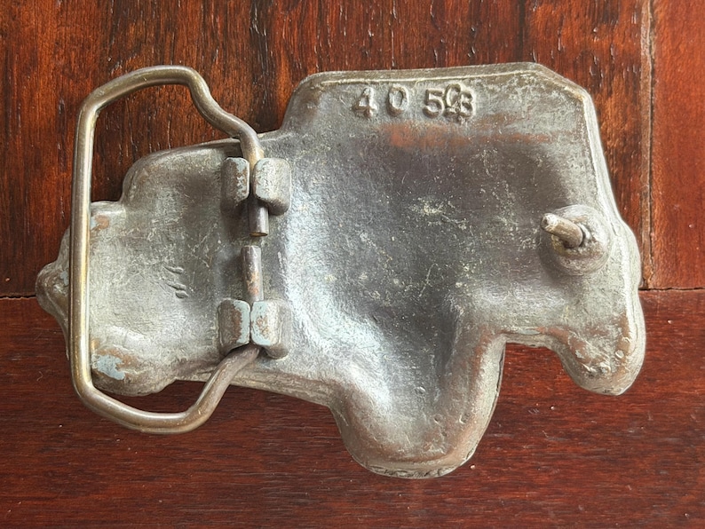 May include: A vintage metal belt buckle with a truck-shaped design. The buckle is a dull silver color with a brass-colored loop and hardware. The numbers "40 503" are stamped on the buckle.