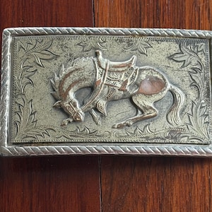 May include: Rectangular silver-toned belt buckle with a detailed relief of a bucking horse wearing a saddle. The horse is set against a textured background with floral and leaf patterns. The buckle has a decorative border.