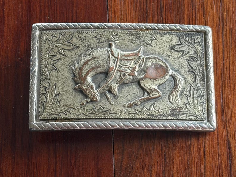 May include: Rectangular silver-toned belt buckle with a detailed relief of a bucking horse wearing a saddle. The horse is set against a textured background with floral and leaf patterns. The buckle has a decorative border.