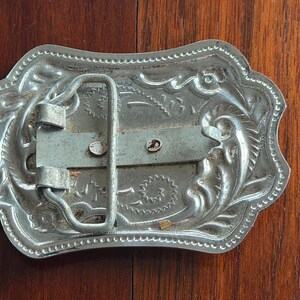 May include: Silver-colored metal belt buckle with an ornate, engraved design. The buckle has a rectangular shape with rounded edges and a decorative border. The back features a metal bar and a loop for the belt.