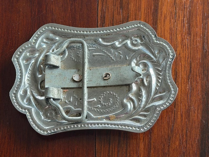 May include: Silver-colored metal belt buckle with an ornate, engraved design. The buckle has a rectangular shape with rounded edges and a decorative border. The back features a metal bar and a loop for the belt.